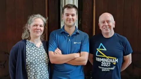 Three people stand side-by-side indoors against a dark wooden wall, facing the camera.

The centre individual wears a blue polo shirt with arms crossed, while the others wear casual tops, including a “Birmingham 2022 Commonwealth Games” T-shirt on the right.

The scene appears warm-lit and informal, with a simple interior setting and no visible decorations beyond wood panelling.