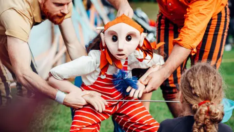 A small human puppet wearing a red and white stripped dungaree and a white shirt with an orange bandana is being controlled by two adults. One man wearing brown with a beard is leaning over. The other person is wearing orange and black stripes. There is a child sat in the foreground watching with a red bow in their long hair.