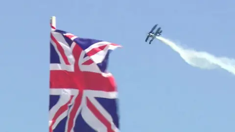 The picture shows a large Union Jack flag in the foreground, waving against a clear blue sky. In the background, a stunt plane is flying at an angle, leaving a long white trail of smoke behind it. The plane appears to be in the middle of an aerobatic manoeuvre, creating a dramatic scene that suggests an airshow or flying display taking place on a bright, sunny day.
