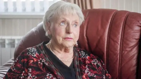 Joanne Coates Head and shoulder view of Ann Ming, an elderly woman with short grey hair. She is wearing a pearl necklace and a jacket with red floral pattern over a black top. Her expression is serious. She is seated on a brown leather sofa, the back of which can be seen behind her.