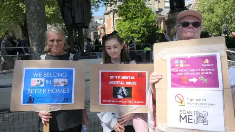 Three woman pose for a photo holding placards protesting about the detention of people with autism. The two either side are around 60 years of age while Lucy, in the centre, is in her twenties. Lucy has long brown hair with sunglasses on her head. 