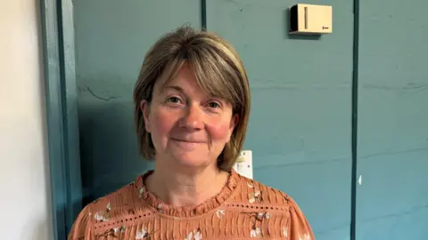 Sarah Gorman stands in front of a blue wall. She is wearing an orange shirt with white flowers on and has short brown hair.