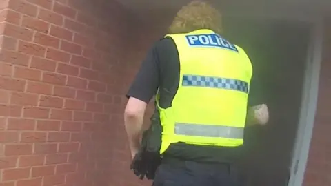 Bodycam still of a police officer stood by an open doorway of a house, with smoke coming from inside