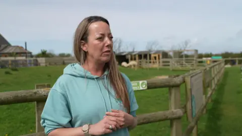 Lisa Vickery is standing next to a grassy enclosure with wooden fencing which has sheep in it. She is talking and holding her hands in front of her. She has long dark blonde hair and is wearing a pale green hoodie. 