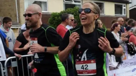 Brain Tumour Research Nick and Rachel Ball running a marathon together. They are both wearing sunglasses and black and green athletic shirts with numbers pinned to them. They are smiling and laughing as they run past some metal barriers where bystanders clap from the side-lines.