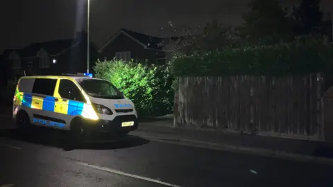 BBC A police vehicle sits beneath a street light next to a dark wood fence and a green shrub.