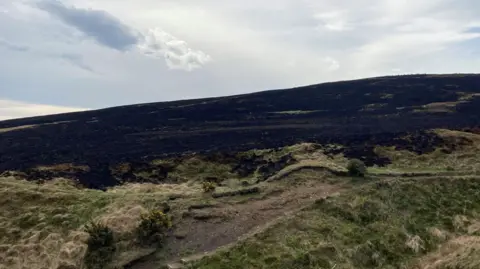 A wide stretch of moorland scarred by fire, with a large area of blackened, scorched ground running across a hillside.