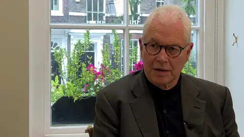 Lord John Hendy, a white haired man with glasses, wearing a grey suit jacket and black shirt