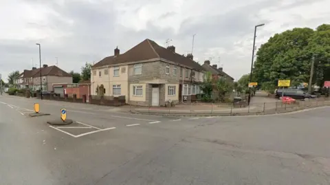 The junction of a small road going on to a main road. There are houses in the top of the photo going of to the right and left. There are also yellow road signs, one with a blue circle and white arrow. Trees and lampposts can also be seen.