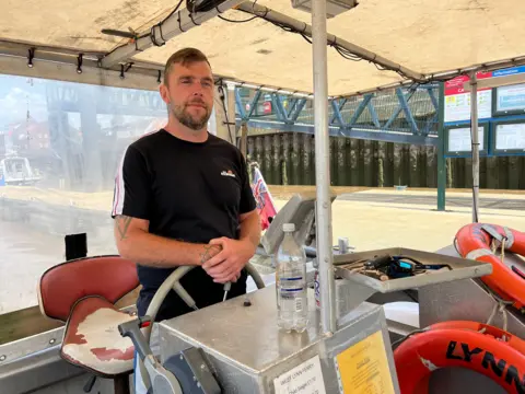 BBC A man standing behind the steering wheel of the boat. He has short brown hair and is wearing a black T-shirt.