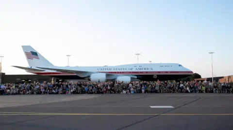 RAF Mildenhall Crowds of people standing and sitting in front of a large white aircraft that has the words "United States of America" on the side and the stars and stripes flag on the tail.