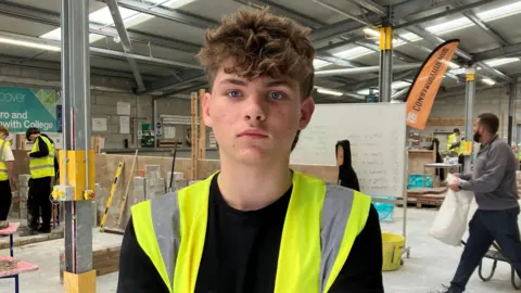 Jack has curly light brown hair over his forehead and wears a high vis yellow jacket and black T-shirt with construction students and equipment behind him. 