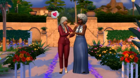 Electronic Arts An in-game screenshot from the game The Sims 4, showing two women walking down the aisle holding each others arms. One woman, with tan skin, in a burgundy suit, has a love heart above her head in a thought bubble. The other woman, who is black with curly grey hair, is wearing a lbue and white dress and smiling.