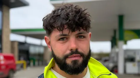 Steve Hubbard/BBC A head and shoulders image of Liam O'Brien. He is wearing a hi-vis top over a white T-shirt and standing on a petrol station forecourt. He is looking directly into the camera.