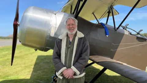 Tim Rayner, a white-haired man with a beard and glasses, stands in front of the plane, wearing a khaki flightsuit and a leather and sheepskin flying jacket. The plane's propeller is on the left of the picture.