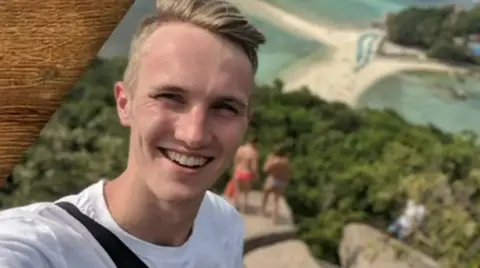 Billy Chip A smiling young man taking a selfie with his back to a lovely scenery of trees and white sand lagoon beaches.