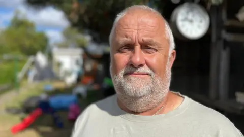 Trevor Dewey looking into camera. He has a white beard and is wearing a light coloured T-shirt. He is in his garden.
