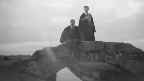 unknown orphan works A black and white photograph of two young men on a small stone bridge over a river or canal, wearing ties with long back robes. One has a pipe in his mouth.