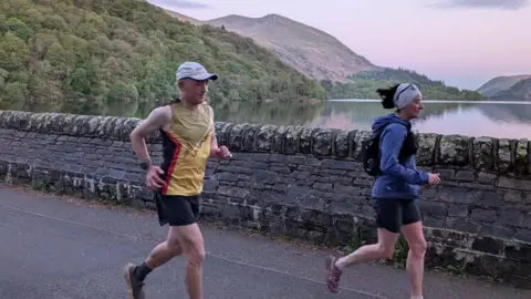 Katy Rodgers Fell runner Kim Collison and another runner are together on a lakeside path in the Lake District. He wears a yellow vest and a beige hate and black shorts and the other runner wears a purple top and black shorts. 