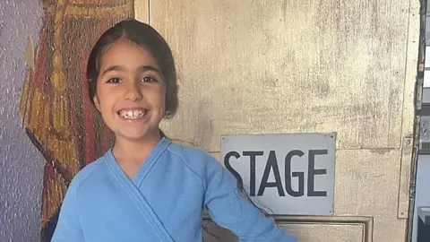 Family photograph The late Alice Aguiar with long black hair in a ponytail wearing a blue ballet top standing in front of a gold door with a sign saying 'stage'. She is smiling. 