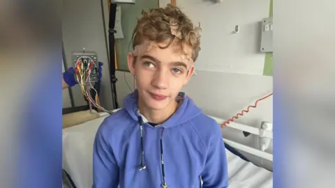 Collect/PA Real Life Tom is wearing a blue jumper, sat on a hospital bed, with wires attached to his head. He is smiling at the camera.