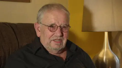 Elton Welsby doing a recent TV interview while sitting on a dark sofa next to a golden-coloured lampshade