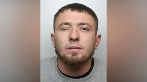 A police mugshot of a man with brown hair and a beard. He is wearing a grey jumper and is standing against a light-coloured backdrop.