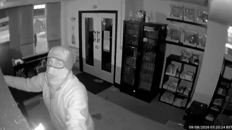 A person wearing a grey balaclava looking into the camera. They appear to be grabbing on to something on a wall.
