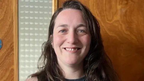 Kelly Quigley has long brown hair and is smiling.
She is standing in front of fire escape door.