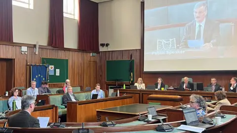 An older man wearing a suit and tie speaks to members of Cheltenham Borough Council in the chamber. He is reading off a sheet of paper and is projected onto a big screen for people to see. Councillors are looking at him.