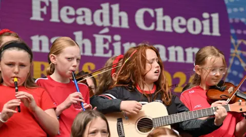A group of young girls play instruments on a stage including a guitar, a fiddle, and tin whistles. They are standing on a stage in front of an Irish language sign which says Fleadh Cheoil na hÉireann.
