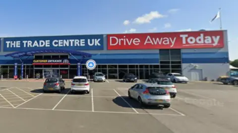 Google Cars are parked in front of a large windowed store front as part of a larger building. There is a blue sign top left with white writing saying "The Trade Centre UK". On the top right of the building is a red sign with white writing saying "Drive Away Today"