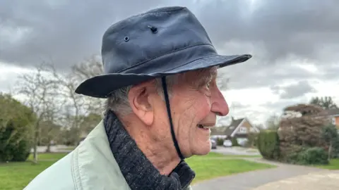 Kimberley Piper/BBC 91-year old Glyn Parry is stood outside facing side-on to the camera. He is wearing a hat with a piece of shoelace tied underneath his chin to keep his hat on.