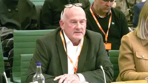 Collier-Keywood speaks to MPs. He is wearing a green blazer and white shirt, and his glasses are perched on top of his head. He has short white hair and is clean shaven. He sits on a green chair with a microphone in front of him, people can be seen sat behind and to the side of him. 