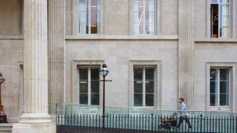 Hawkins Brown A ramp with glass panels along the side of it leads up to a grand building with neoclassical pillars, with a man pushing a pram walking up it