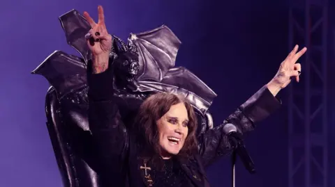 A performer seated on a black throne with a bat-shaped backrest raises both arms in a “peace” sign gesture while speaking into a microphone on a stage lit with purple lighting.
