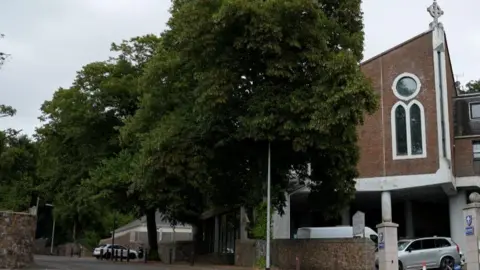 Tom Drysdale/BBC Two tall trees are in leaf in front of a brick covered church with a round and an arched church window and a white stone cross on the top of the sloping roof.
