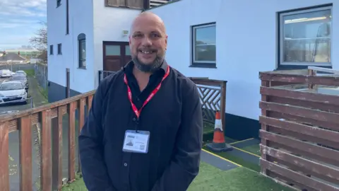 Justin Bellinger in a black shirt. He is wearing a red lanyard. Behind him is a white building and a car park. 