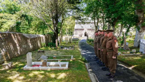Lucie Stribrska The image shows a group of six soldiers standing in a line on a paved path within a cemetery. They are dressed in historical military uniforms from the World War II era, with brown tunics, trousers, and caps.