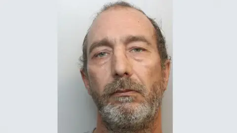 Wiltshire Police A police mugshot of Tommy Hitt. He has green eyes, short brown hair which is balding on the top and a scruffy grey beard. He is looking at the camera with a blank expression.