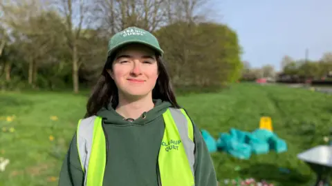 A girl with long black hair is smiling and looking at the camera. She is wearing a green hat and hoodie as well as a high-vis vest, all adorned with Cleanup Quest.