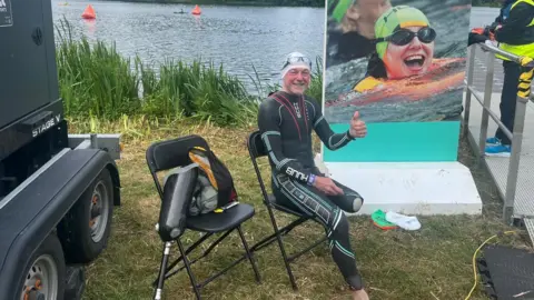 Chris and Denise Arthey A man with one leg sat by a lake in a wetsuit and swim hat and goggles. He is smiling with his thumb up. 