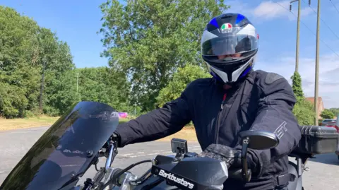 Lee is sitting on his motorbike with a blue, black and white helmet dressed in a black jacket and gloves on a black bike
