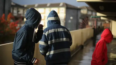 Three teenage boys wearing hoodies. They are standing outside a block of flats. The concrete walkway is wet as if it has been raining recently.