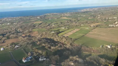 BBC An aerial view of Guernsey from above, mainly fields and trees, with buildings sparsely visible. Blue skies and ocean seen from afar.