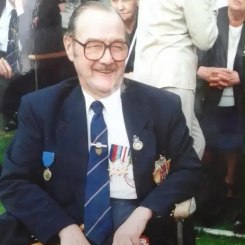 Family handout The photo and shows a man smiling to the right of the camera. He is sat down, and has thick black glasses. He has a navy blazer, as well as a shirt and tie. He also has medals attached to the right hand side of his blazer. 