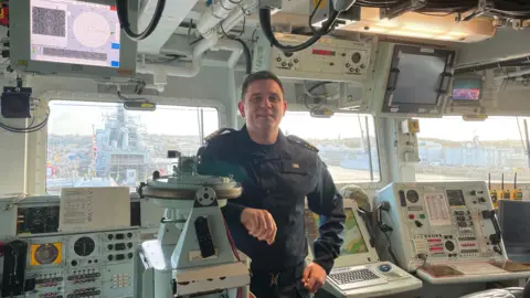 Executive Officer Byron Linn leaning on the compass on the bridge of HMS St Albans in a dark blue uniform. 