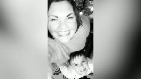 A woman smiles up at the camera. She is taking a selfie with a young boy who is also smiling at the camera. The photo is in black and white.