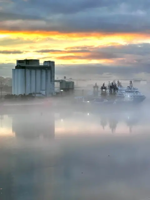 Isobel Irvine Industrial harbor scene at sunrise with silos and a docked ship partially obscured by thick fog, reflecting on calm water.”