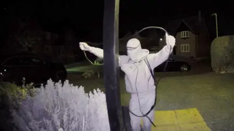 Man wearing a balaclava holding up a relay device while stealing a keyless car.